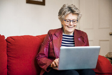 Ältere Frau auf dem Wohnzimmersofa beim Tippen auf dem Laptop - CUF20278