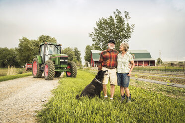 Paar mit Hund auf einem Bauernhof vor einem Traktor, das lächelnd in die Kamera schaut - CUF20330