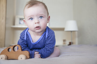 Porträt eines blauäugigen kleinen Jungen, der mit einem Holzspielzeugauto auf dem Bett sitzt - CUF20390