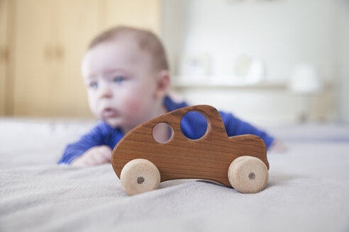 Kleiner Junge spielt auf dem Bett mit einem Spielzeugauto aus Holz - CUF20391