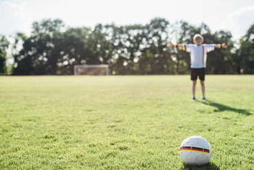 Junge mit ausgestreckten Armen steht auf einem Fußballfeld zwischen einem deutschen Fußball und einem Tor - MJF02307