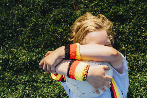 Boy in German soccer shirt lying on grass, hugging ball - MJF02312