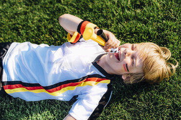 Boy in German soccer shirt lying on grass, blowing horns - MJF02316