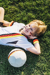 Junge in deutschem Fußballtrikot auf dem Rasen liegend, lächelnd - MJF02339