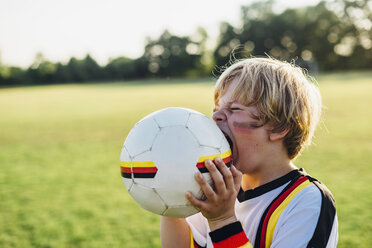 Junge mit Gesichtsbemalung und deutschem Fußballtrikot, der in einen Fußball beißt - MJF02342