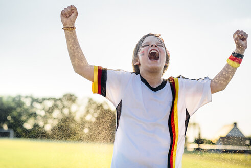 Boy wearing German soccer shirt screaming for joy, standing in water splashes - MJF02347
