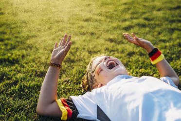 Junge in deutschem Fußballtrikot auf dem Rasen liegend, lachend und schreiend - MJF02350