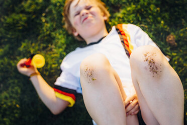 Boy in German soccer shirt lying on grass, with dirty knees - MJF02354