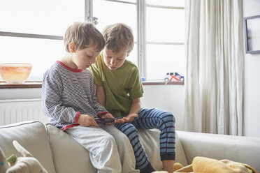 Jungen im Schlafanzug sitzen auf dem Sofa und schauen auf ihr Smartphone - CUF20604