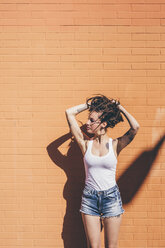 Young woman with hands in dreadlock hair in front of orange wall - CUF20834