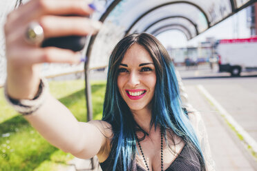 Junge Frau mit gefärbten blauen Haaren, die in einem Wartehäuschen ihr Smartphone aufnimmt - CUF20841