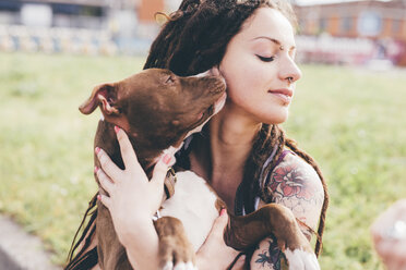 Pit bull terrier licking tattooed young woman in urban park - CUF20845