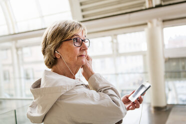 Seitenansicht einer älteren Frau, die In-Ear-Kopfhörer einstellt, während sie mit ihrem Smartphone am Bahnhof steht - MASF07611