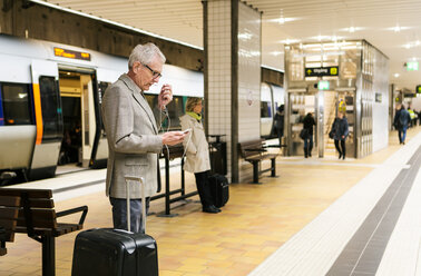 Älterer männlicher Pendler, der sein Smartphone benutzt, während er neben seinem Gepäck in der U-Bahn-Station steht - MASF07620