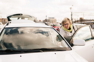 Senior woman standing by car in parking lot against sky - MASF07624