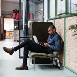 Full length of young businessman using laptop while sitting on chair at creative office - MASF07656