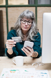 Ältere Geschäftsfrau, die mit ihrem Mobiltelefon online einkauft, während sie im Büro einen Kaffee trinkt - MASF07671