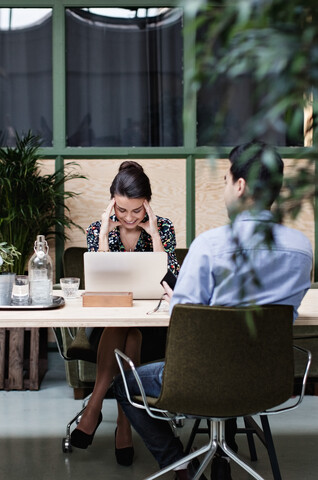 Müde Geschäftsfrau, die einen Laptop benutzt, während sie mit einem Kollegen am Tisch sitzt, lizenzfreies Stockfoto