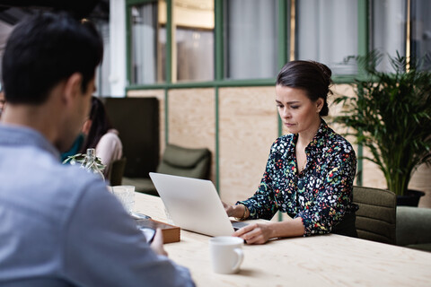 Geschäftsfrau, die einen Laptop benutzt, während sie mit einem Kollegen an einem Tisch im Büro sitzt, lizenzfreies Stockfoto