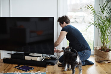 Schwangere Frau ordnet die Kabel des Fernsehers, während sie neben dem Hund im Wohnzimmer kniet - MASF07711