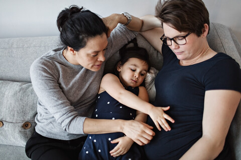 High angle view of mother and daughter touching woman's abdomen while leaning on sofa at home stock photo