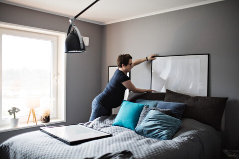 Schwangere Frau arrangiert Bilder auf grauer Wand im Schlafzimmer zu Hause, lizenzfreies Stockfoto