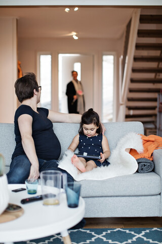 Mother sitting with daughter on sofa looking at woman entering house stock photo