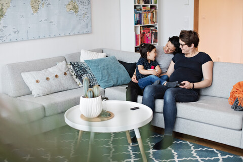 Lächelndes lesbisches Paar sitzt mit Tochter auf dem Sofa im Wohnzimmer zu Hause, lizenzfreies Stockfoto