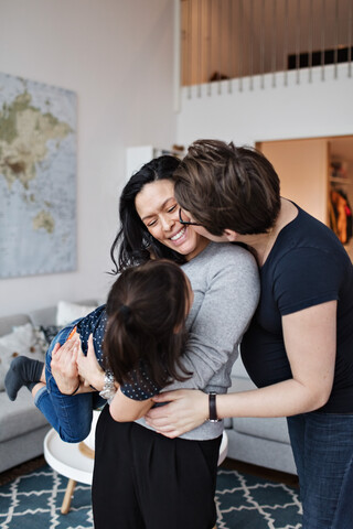 Frau küsst Freundin mit Tochter im Wohnzimmer zu Hause, lizenzfreies Stockfoto
