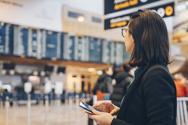 Mittlere erwachsene Geschäftsfrau, die ein Mobiltelefon hält, während sie im Flughafen steht - MASF07800