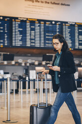 Businesswoman using mobile phone while walking with luggage in airport terminal - MASF07803