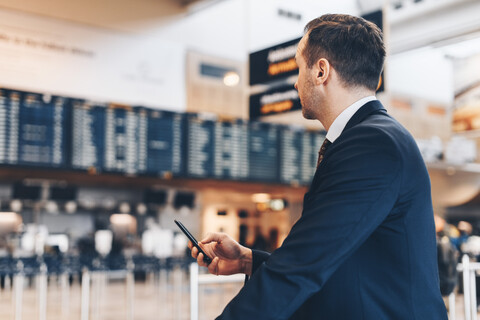 Geschäftsmann, der ein Mobiltelefon benutzt, während er im Flughafenterminal wegschaut, lizenzfreies Stockfoto