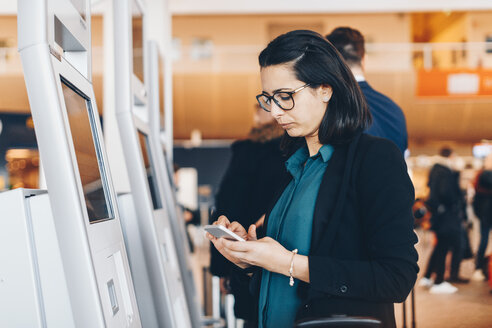 Geschäftsfrau, die ein Mobiltelefon am automatischen Check-in-Automaten im Flughafen benutzt - MASF07807