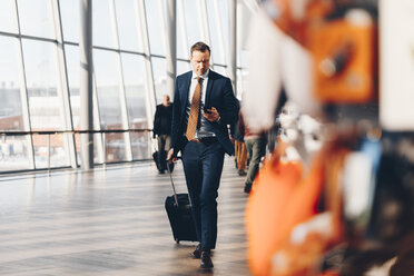 Reifer Geschäftsmann mit Gepäck in voller Länge, der beim Gehen im Flughafen-Terminal ein Mobiltelefon benutzt - MASF07809