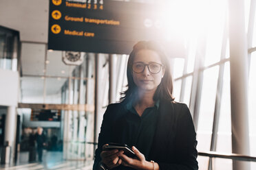 Portrait of confident businesswoman holding mobile phone and passport in airport terminal - MASF07822