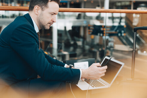 Seitenansicht eines Geschäftsmannes, der ein Mobiltelefon und einen Laptop im Flughafen benutzt - MASF07837