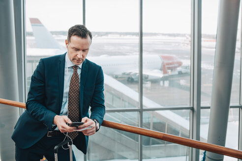 Geschäftsmann, der ein Mobiltelefon benutzt, während er sich am Geländer eines Flughafens abstützt - MASF07846