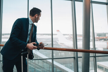 Businessman looking at airplane through window while using mobile phone in airport - MASF07847