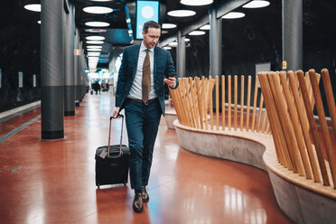 Full length of businessman with luggage and mobile phone walking in railroad platform - MASF07852