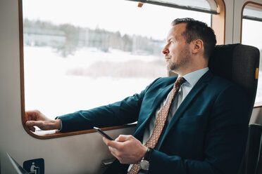 Nachdenklicher Geschäftsmann, der sein Mobiltelefon während einer Zugfahrt in der Hand hält - MASF07858
