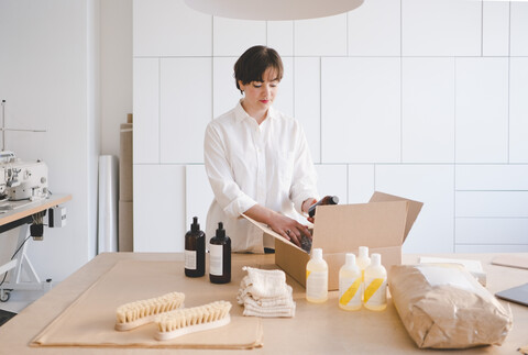 Unternehmerin beim Auspacken eines Kartons mit Flaschen am Tisch in der Werkstatt, lizenzfreies Stockfoto