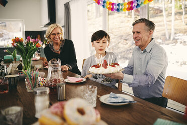 Junge bläst Kerzen auf Geburtstagskuchen aus, während er mit seinen Großeltern am Tisch sitzt - MASF07929