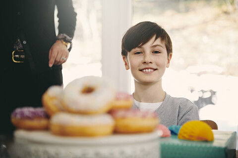 Porträt eines lächelnden Jungen, der bei seiner Großmutter am Esstisch sitzt, mit Donuts im Vordergrund, lizenzfreies Stockfoto