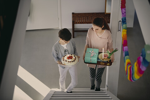 Hochformatige Ansicht von Geschwistern, die eine Geburtstagstorte und ein Geschenkpaket tragen, während sie zu Hause die Treppe hinaufsteigen, lizenzfreies Stockfoto