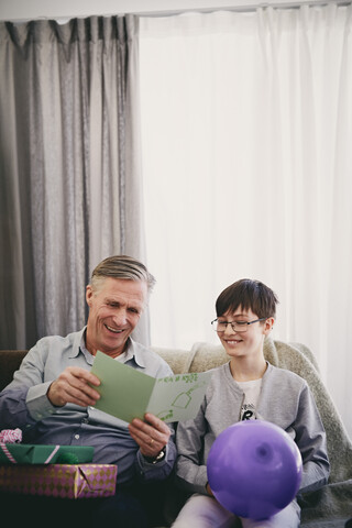 Lächelnder Großvater liest eine Grußkarte, während er mit seinem Enkel und Geschenken auf einer Party im Wohnzimmer sitzt, lizenzfreies Stockfoto