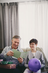 Lächelnder Großvater liest eine Grußkarte, während er mit seinem Enkel und Geschenken auf einer Party im Wohnzimmer sitzt - MASF07949
