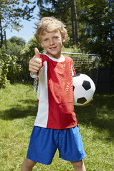 Porträt eines Jungen in Fußballtrikot, der einen Fußball hält und im Garten die Daumen nach oben streckt - CUF21000