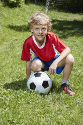 Porträt eines Jungen in Fußballkleidung, der mit einem Fußball im Garten hockt - CUF21001