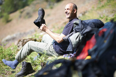 Mature man sitting on hillside holding hiking boot smiling - CUF21029