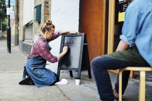 Junge Frau kniet auf dem Bürgersteig und schreibt auf einer Tafel vor einer Werkstatt - CUF21076